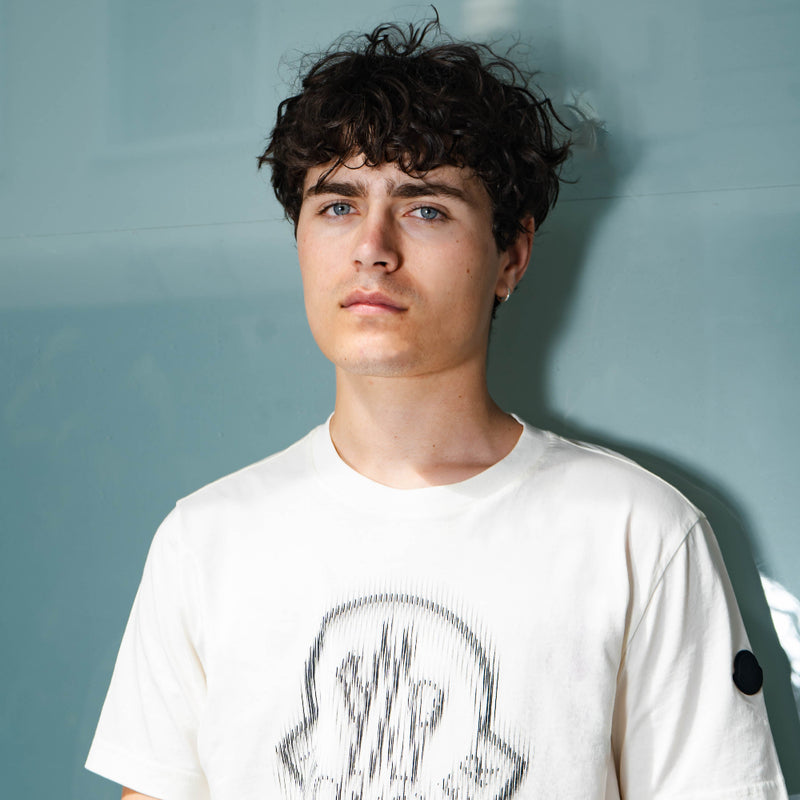 Person wearing a white Moncler  t-shirt with a graphic design against a light blue wall
