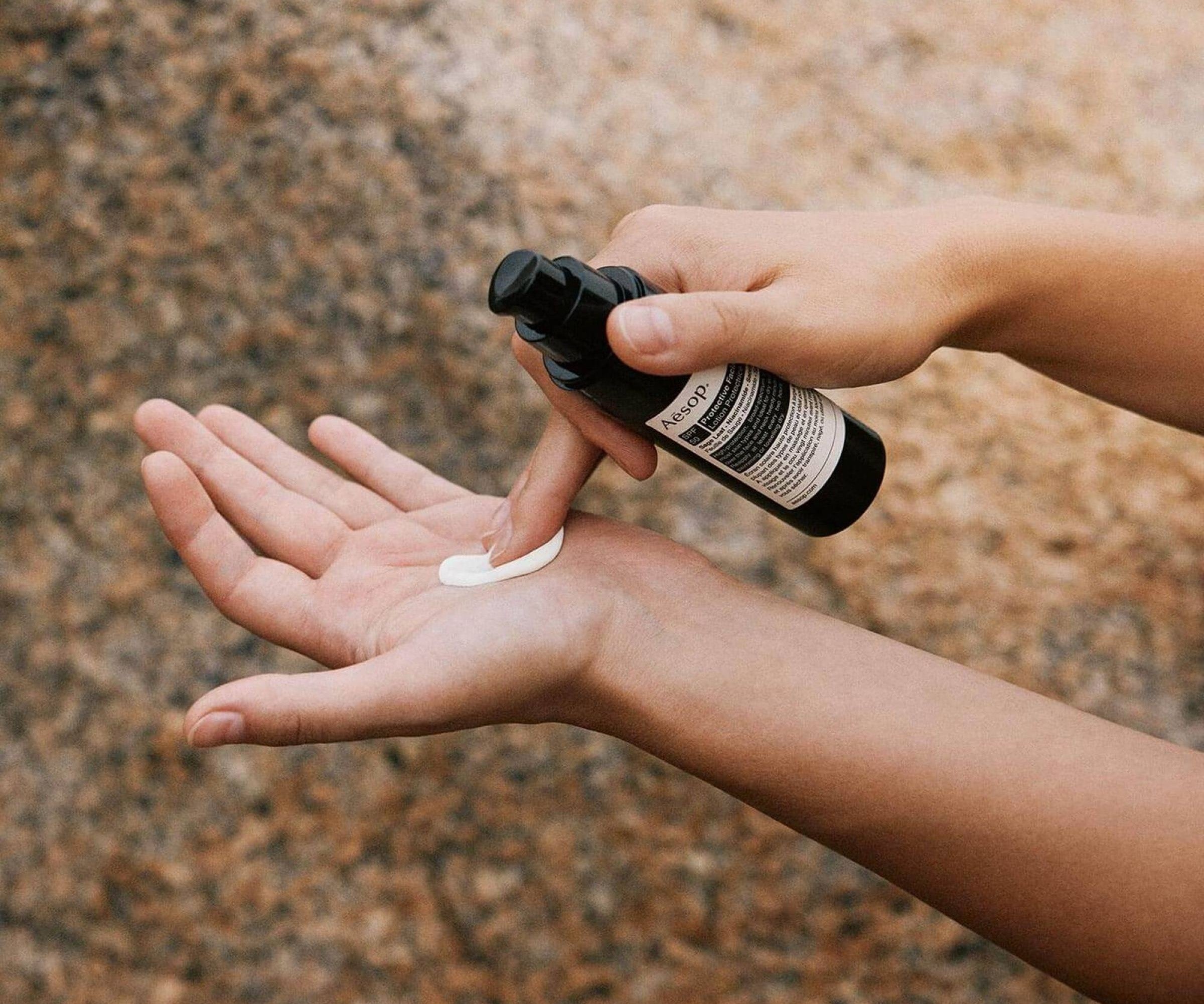 Person applying a white cream from a black bottle onto their palm against a brown stone background