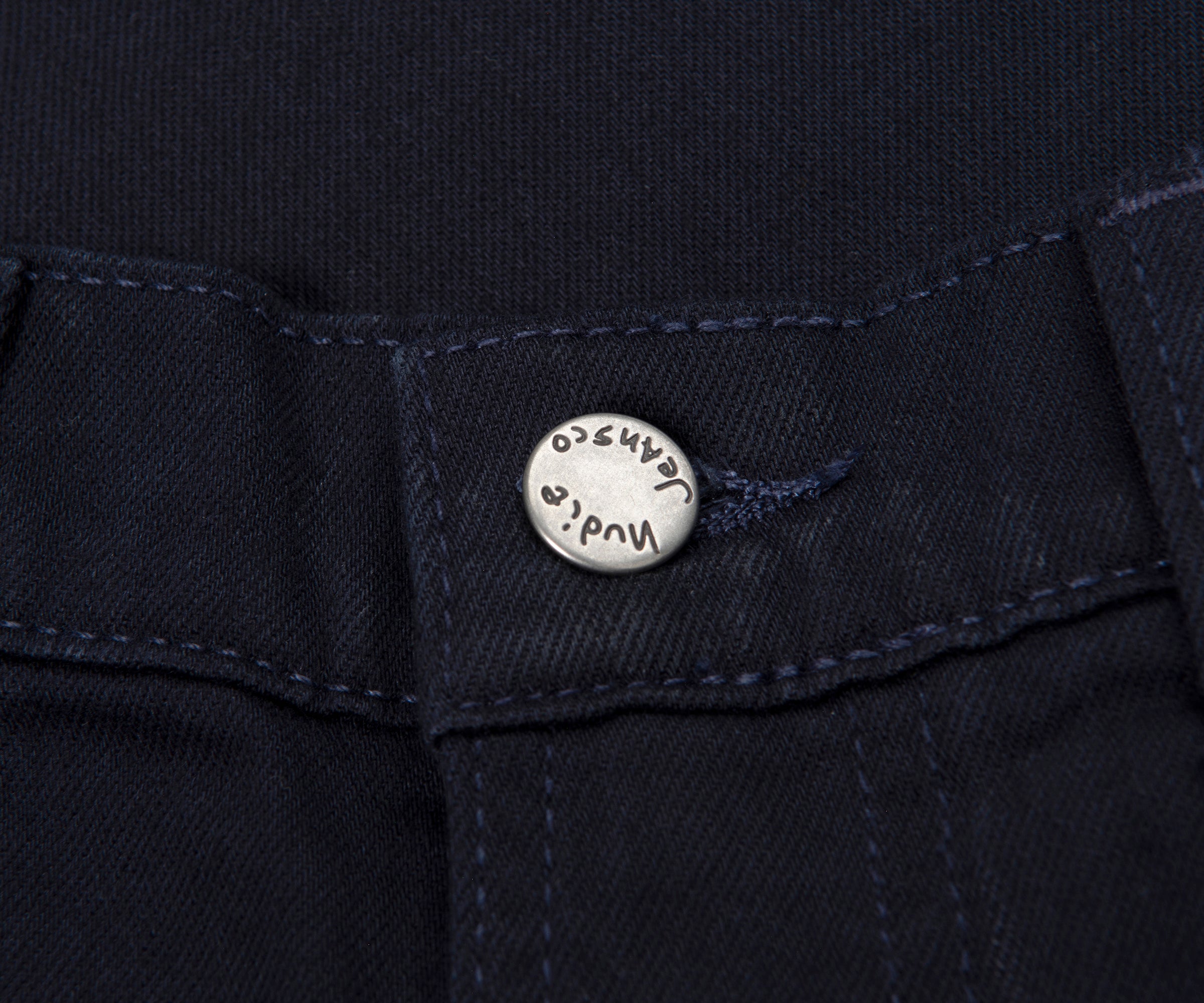 Close-up of a button on a dark fabric, likely jeans, with 'Nudie Jeans' branding.