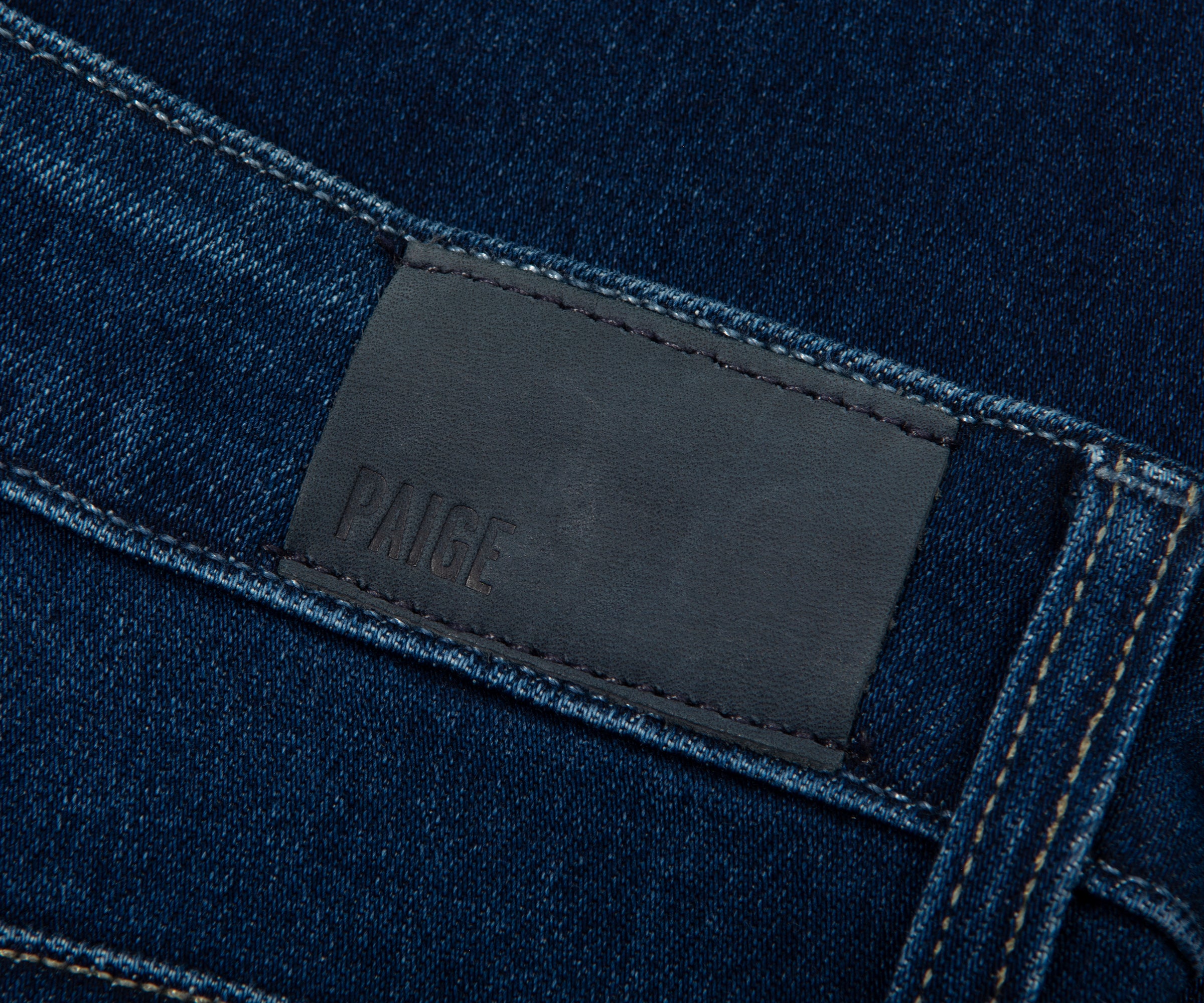 Close-up of a dark blue denim fabric with a black leather label featuring the brand 'PAIGE'.