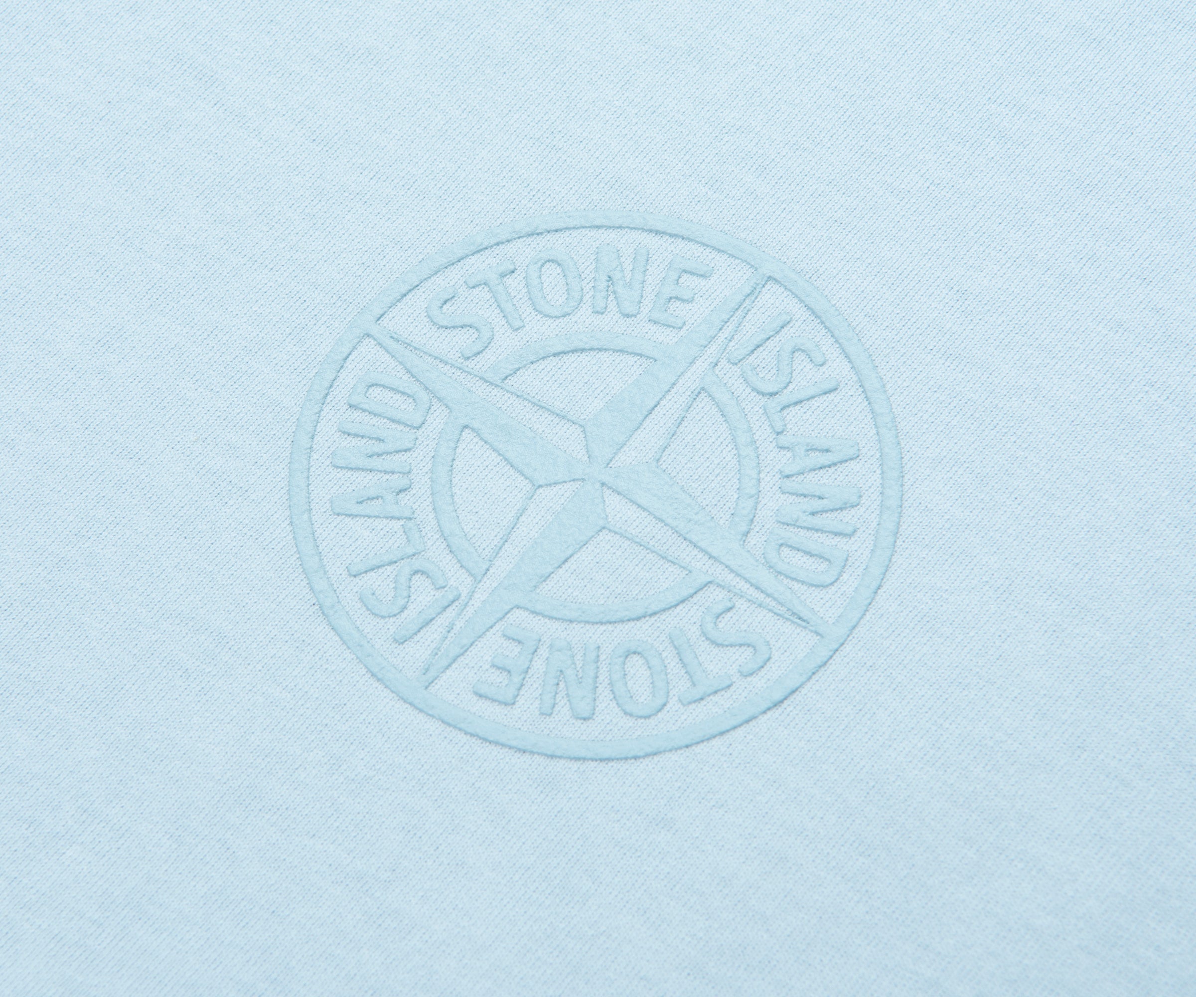 Embossed Stone Island logo on a light blue textured fabric background